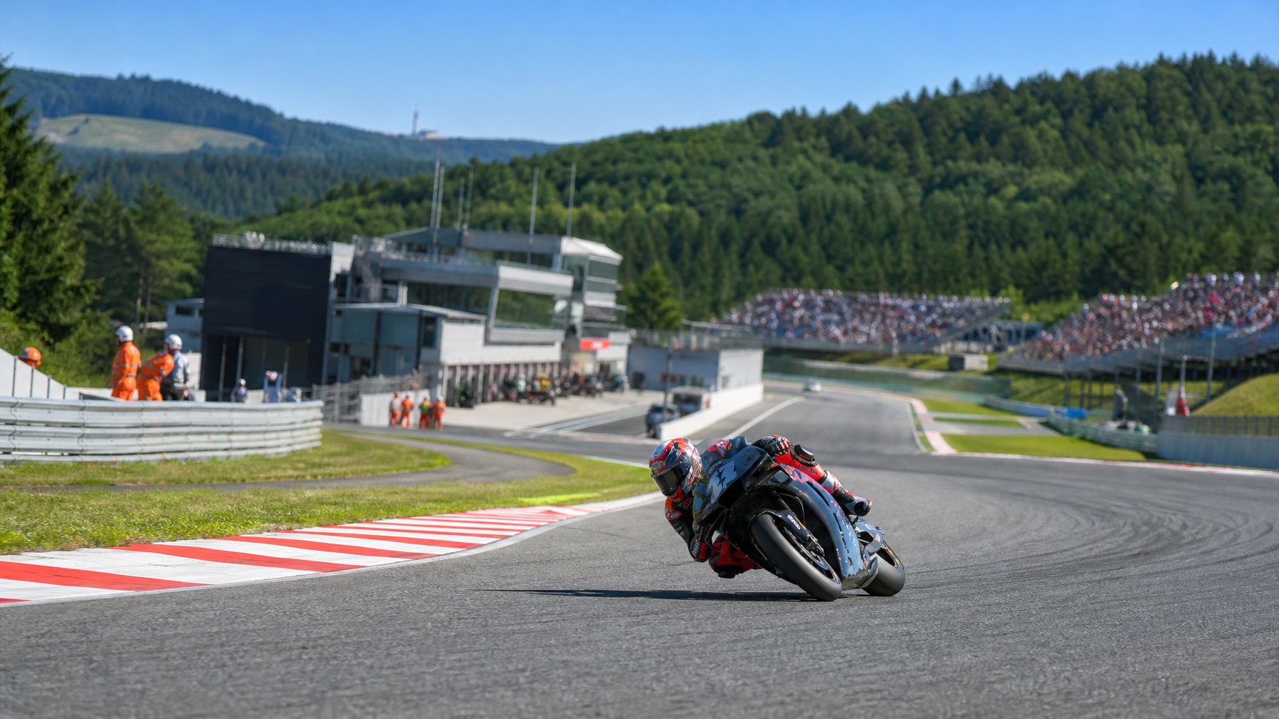Pembalap MotoGP melaju di tikungan Sirkuit Brno dengan latar perbukitan hijau dan tribun penonton.
