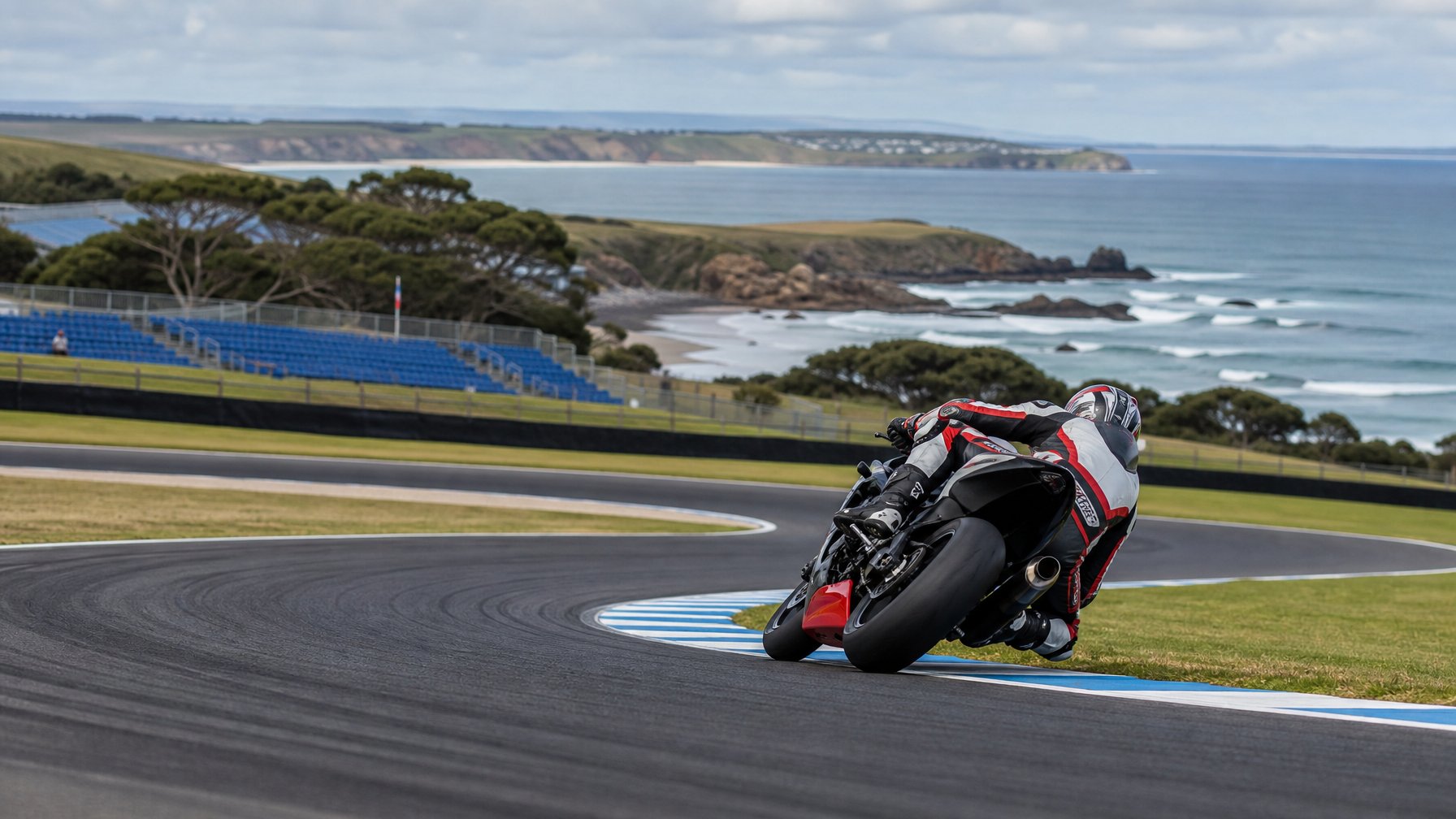 Pembalap motor menikung tajam di Sirkuit Phillip Island dengan garis pantai Australia di latar belakang.