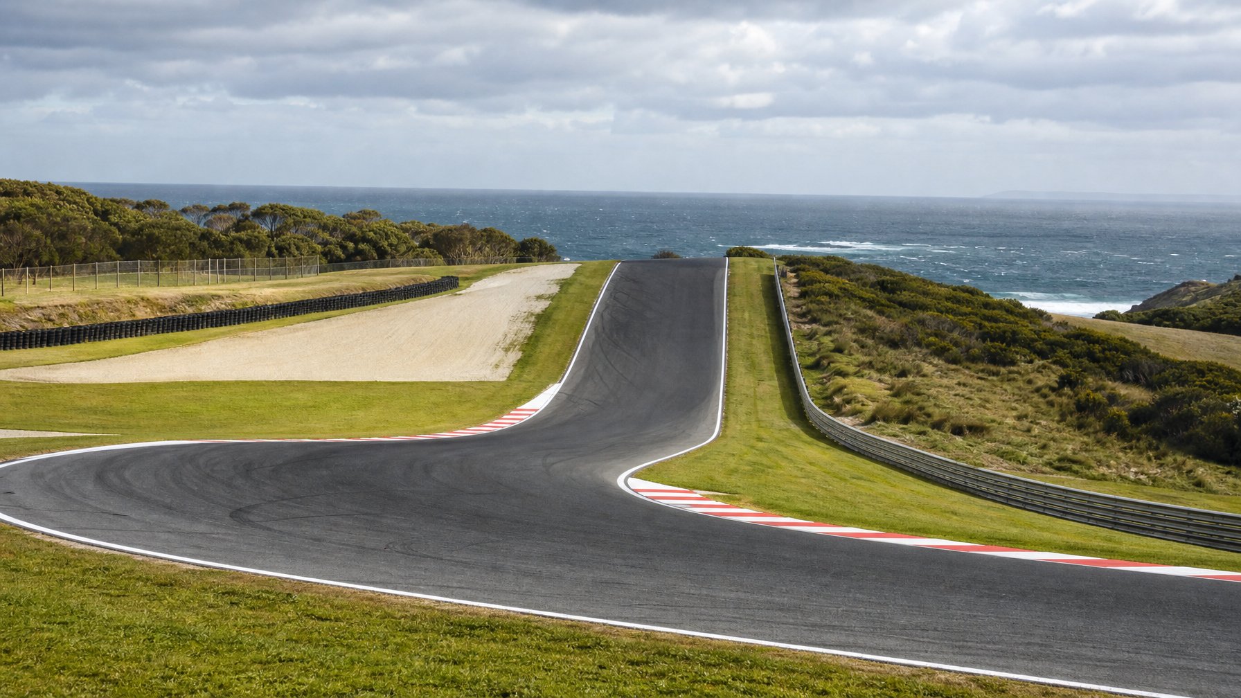 Tikungan menantang di Sirkuit Phillip Island dengan perubahan elevasi, kerb balap, area rumput, dan garis pantai di kejauhan.