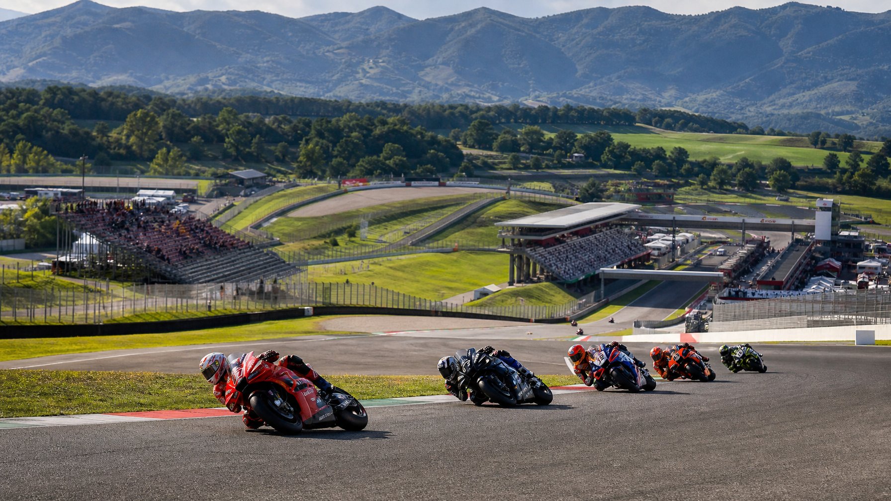 Pembalap MotoGP melaju di tikungan Sirkuit Mugello dengan latar perbukitan hijau Tuscany.