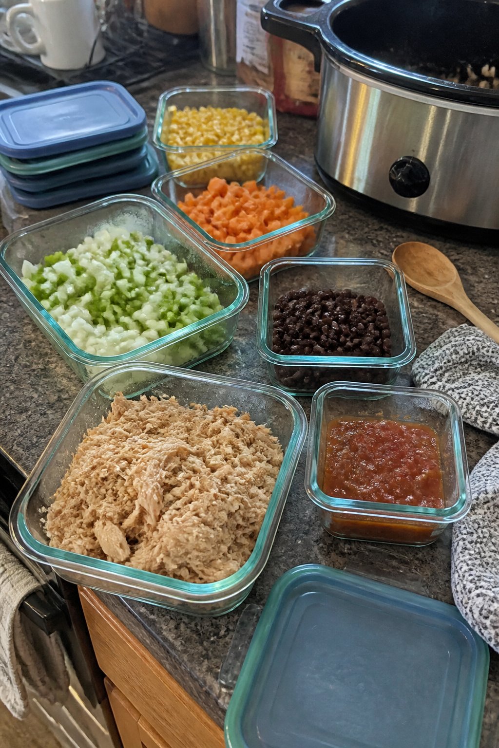 12 Crockpot Accessories That Make Dump Dinners Easier 10 Glass food storage containers holding prepared ingredients sit beside a slow cooker on a kitchen counter.