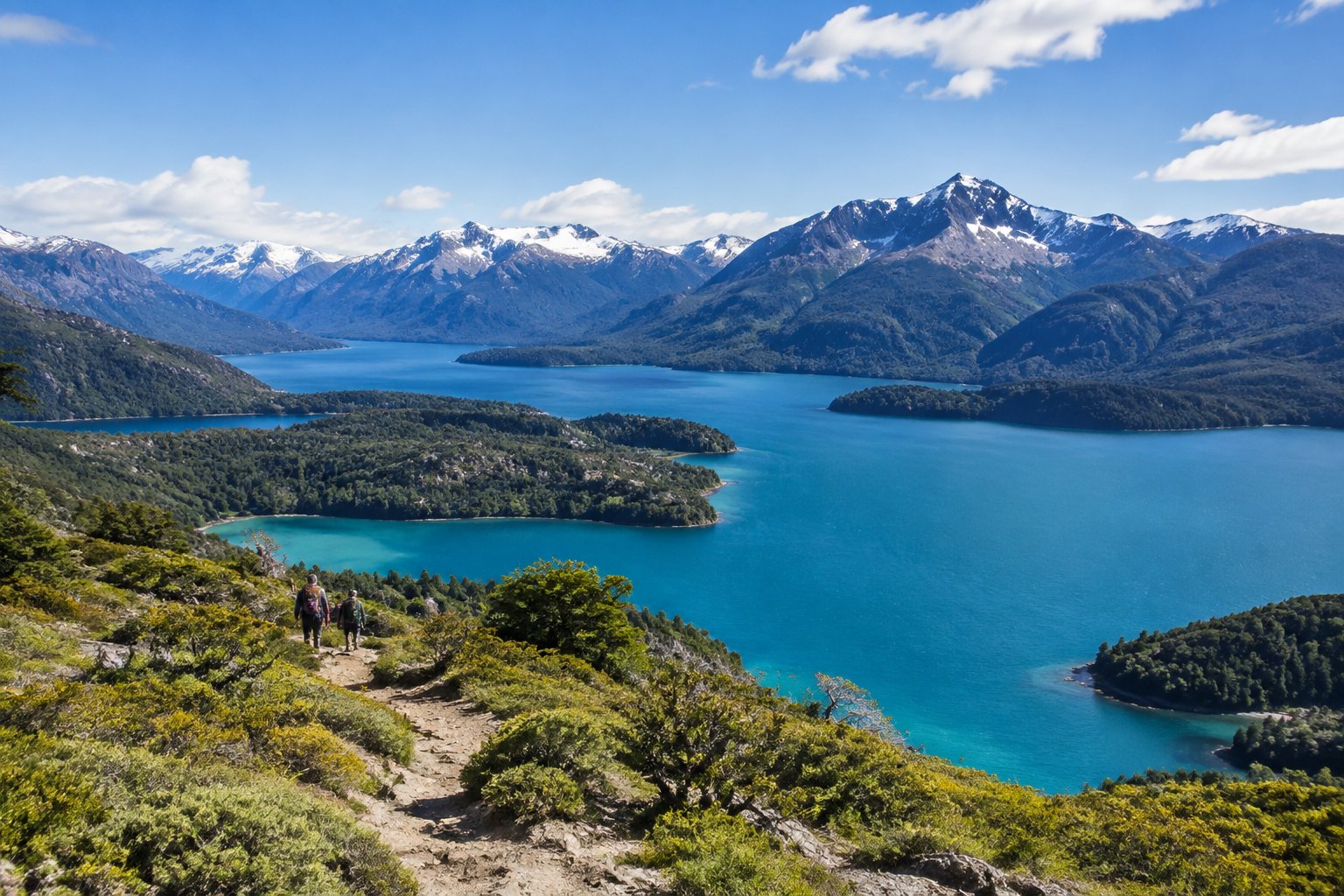 Hikers overlook a turquoise Patagonian lake surrounded by forested mountains.