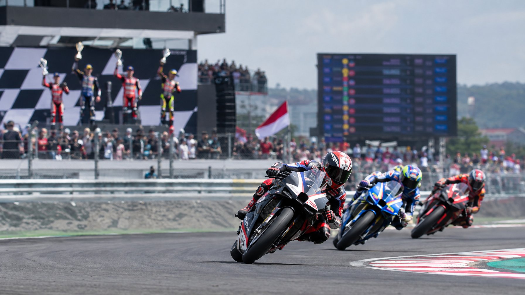Pembalap MotoGP bersaing di tikungan sirkuit, dengan perayaan podium dan bendera Indonesia di latar belakang.
