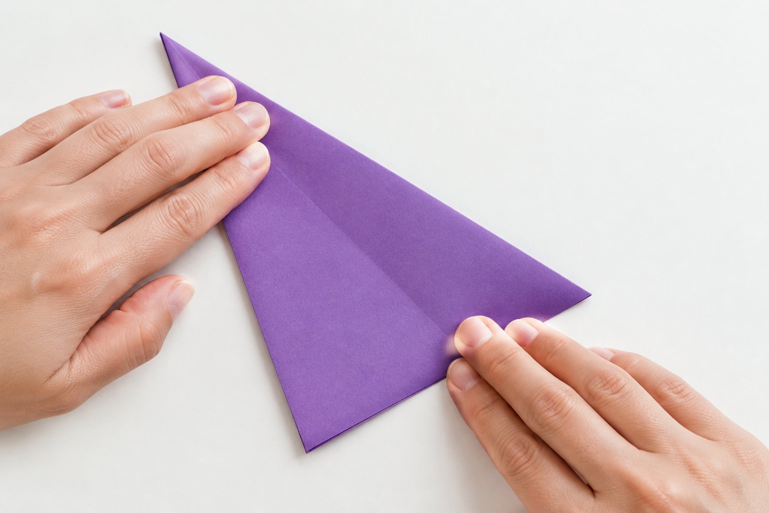 Hands fold a square sheet of purple paper diagonally on a table.