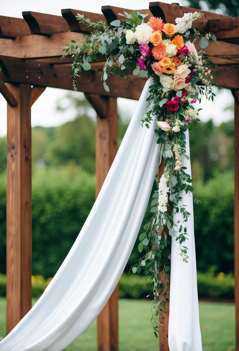 A white satin drape cascades over a wooden wedding pergola, adorned with vibrant fresh flowers in various hues