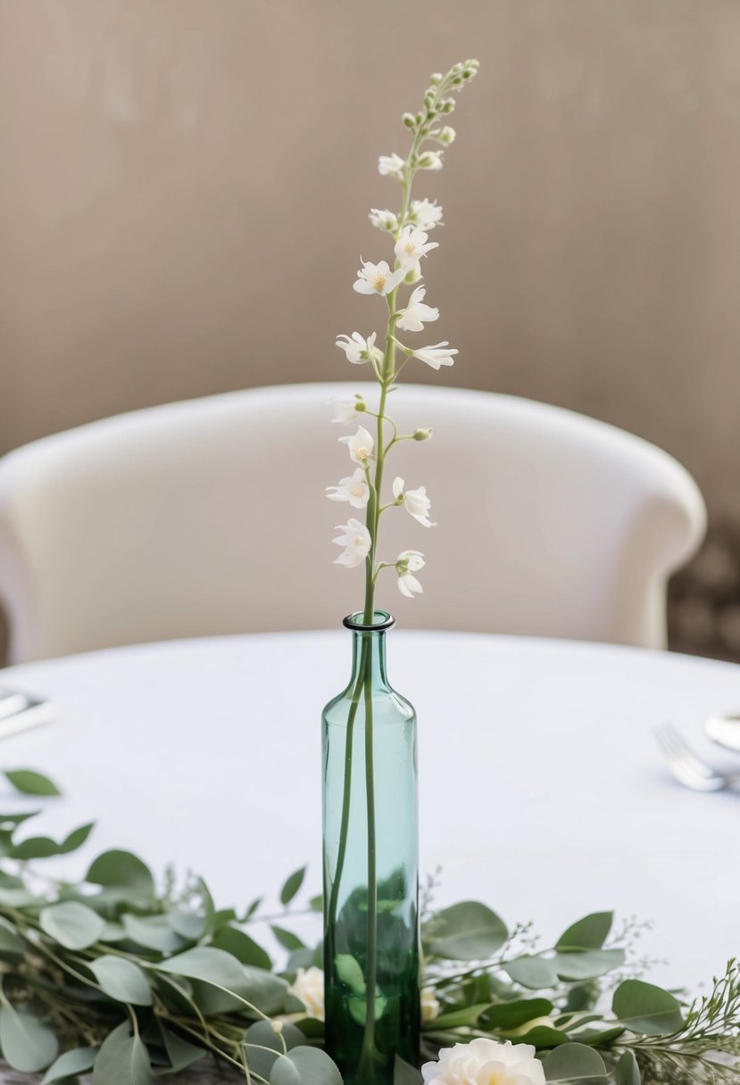 A long, slender vase holds a single, delicate flower stem, surrounded by soft greenery on a simple, bohemian-inspired wedding table