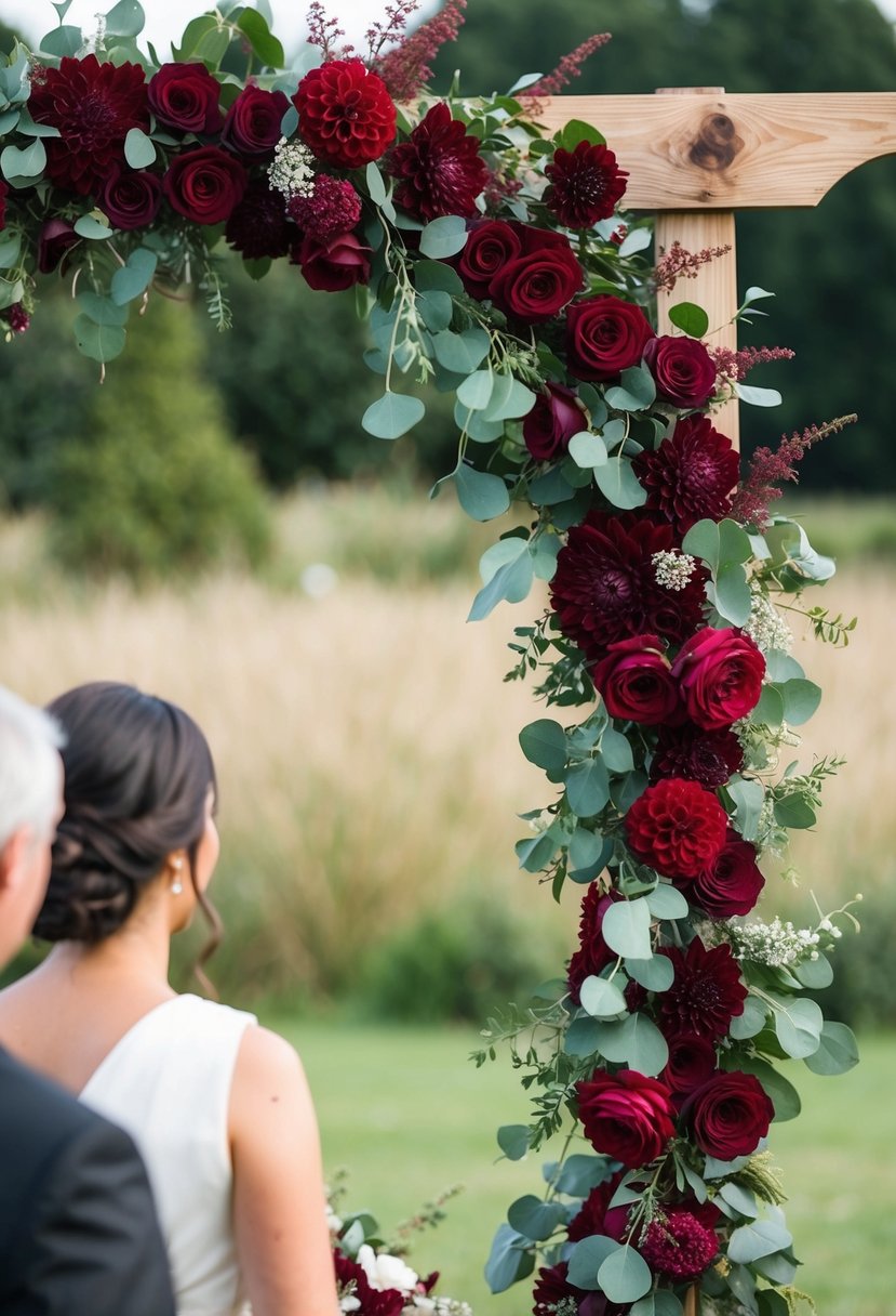 A lush burgundy floral arch frames a rustic outdoor wedding ceremony. Deep red roses, dahlias, and eucalyptus adorn the wooden structure